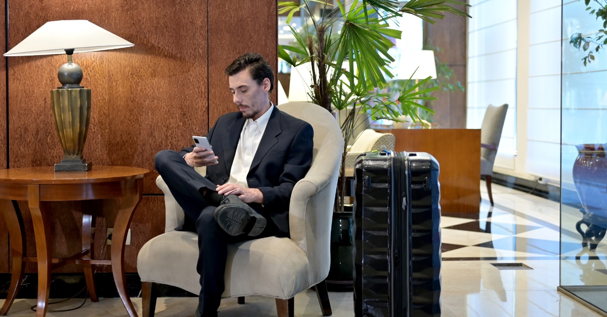 Man sitting on a chair in a hotel lobby looking on his phone, with a big suitcase next to him