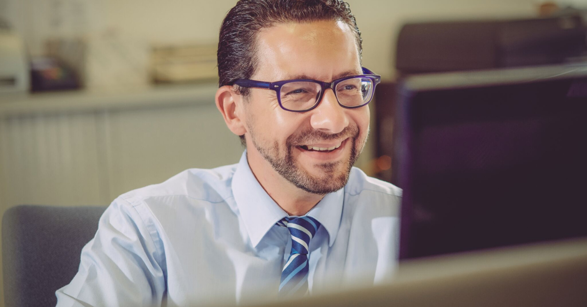 A man in a shirt and tie wearing glasses and smiling at a computer screen